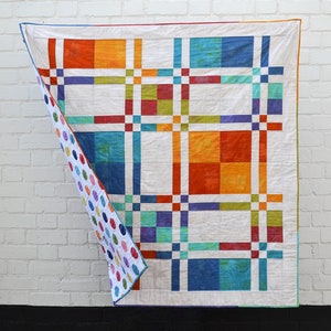 Könnte beinhalten: Ein farbenfroher Quilt mit einem Gittermuster aus orangefarbenen, blauen, grünen und lila Quadraten auf weißem Hintergrund. Die Rückseite des Quilts zeigt ein Muster aus bunten Punkten. Der Quilt hängt vor einer weißen Backsteinmauer.