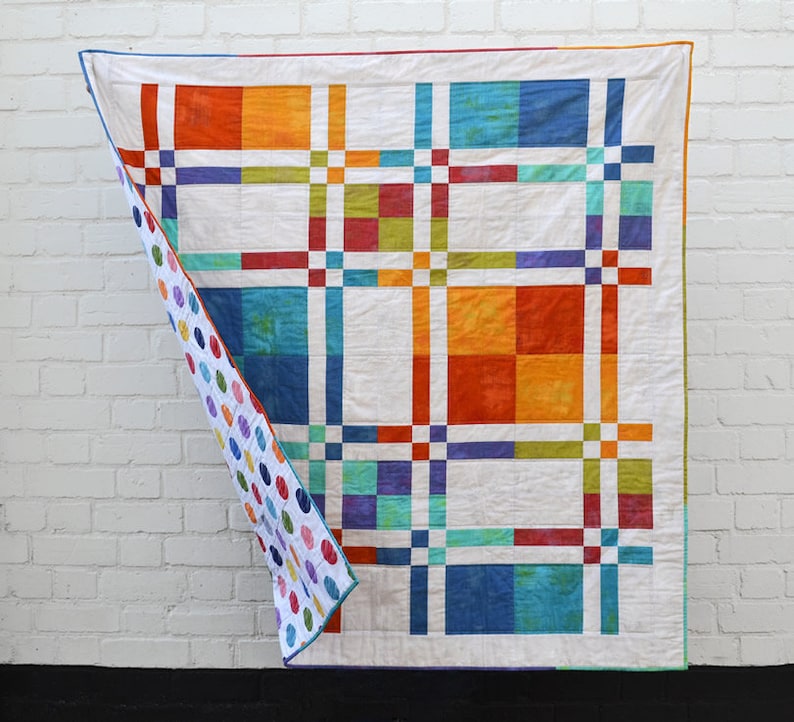 K&ouml;nnte beinhalten: Ein farbenfroher Quilt mit einem Gittermuster aus orangefarbenen, blauen, gr&uuml;nen und lila Quadraten auf wei&szlig;em Hintergrund. Die R&uuml;ckseite des Quilts zeigt ein Muster aus bunten Punkten. Der Quilt h&auml;ngt vor einer wei&szlig;en Backsteinmauer.
