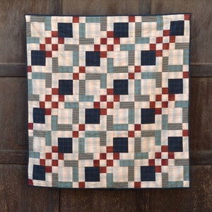 May include: A handmade quilt with a geometric pattern. The design features alternating squares of navy blue, red, and light blue, set against a cream-colored background with plaid accents. The quilt is displayed against a dark wood background.