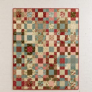 Può includere: Una trapunta patchwork rettangolare caratterizzata da una griglia di blocchi di tessuto quadrati con vari motivi e colori, tra cui tonalità di rosso, blu, verde e crema. La trapunta è rifinita con un bordo in tessuto rosso tinta unita sui quattro lati.