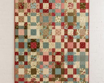May include: A rectangular patchwork quilt featuring a grid of square fabric blocks in various patterns and colours including deep red, muted blue, cream, and olive green. The quilt is finished with a solid red fabric border along all four edges.
