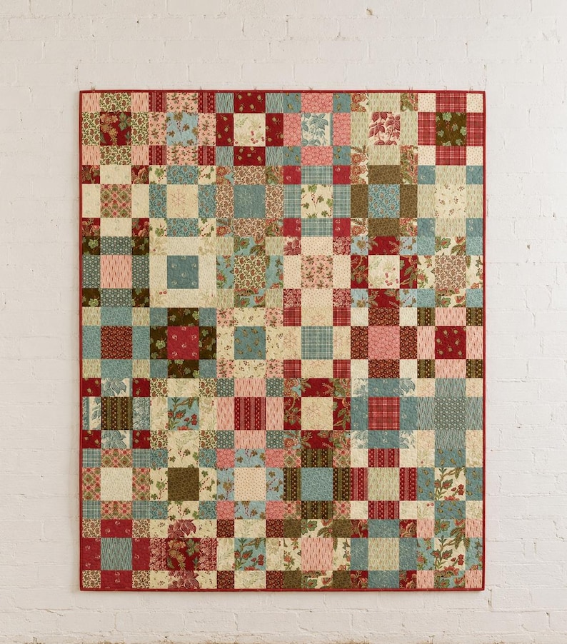 May include: A rectangular patchwork quilt featuring a grid of square fabric blocks in various patterns and colours, including shades of red, blue, green, and cream. The quilt is finished with a solid red fabric border along all four edges.