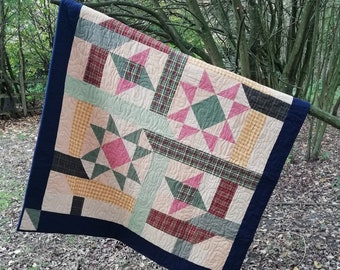 May include: A handmade quilt with a navy blue border, displayed outdoors. The quilt features a patchwork design with star patterns in pink, green, and red plaid, set against a beige background. The quilt is hanging from a tree branch.