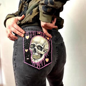 May include: Black denim jeans with a painted skull patch on the back pocket. The skull is white with pink dripping paint and yellow hearts around it.