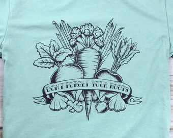 May include: A light blue t-shirt featuring a black and white illustration of various root vegetables, including carrots, beets, and radishes. The design includes the text "DON'T FORGET YOUR ROOTS" on a banner. The shirt is made of a soft material.