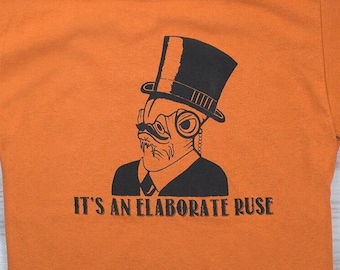 May include: Orange t-shirt with a black and white illustration of a fish wearing a top hat and monocle. The text "It's an elaborate ruse" is printed below the illustration.