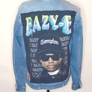 May include: Light blue denim jacket with a black graphic on the back featuring the text "EAZY-E" in a 3D font and a portrait of a man wearing a hat that says "Compton". The jacket has a classic collar and button closure.