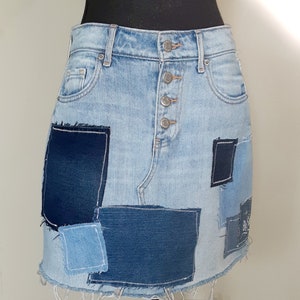May include: A light blue denim mini skirt with a patchwork design. The skirt has multiple squares of different shades of blue denim sewn onto the front. The skirt has a button fly and a frayed hem.