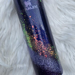 May include: A stainless steel tumbler with a dark blue base, adorned with colorful glitter and a blue lid. The Stanley logo is visible on the side. The tumbler is designed to keep beverages hot or cold.