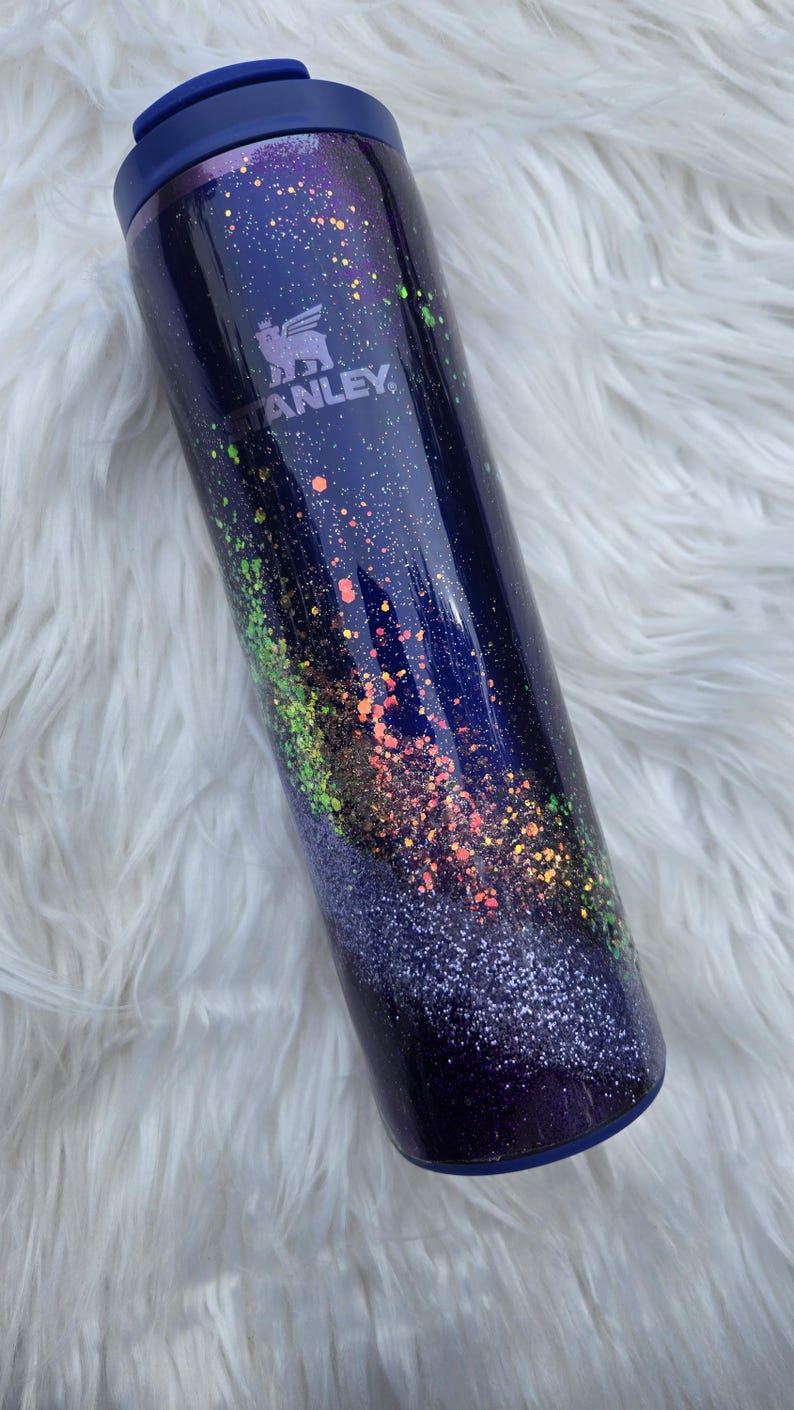 May include: A stainless steel tumbler with a dark blue base, adorned with colorful glitter and a blue lid. The Stanley logo is visible on the side. The tumbler is designed to keep beverages hot or cold.
