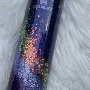 May include: A stainless steel Stanley tumbler with a navy blue base and lid. The tumbler is decorated with a galaxy-themed design, featuring colorful speckles of orange, green, and purple glitter. The Stanley logo is visible on the side.