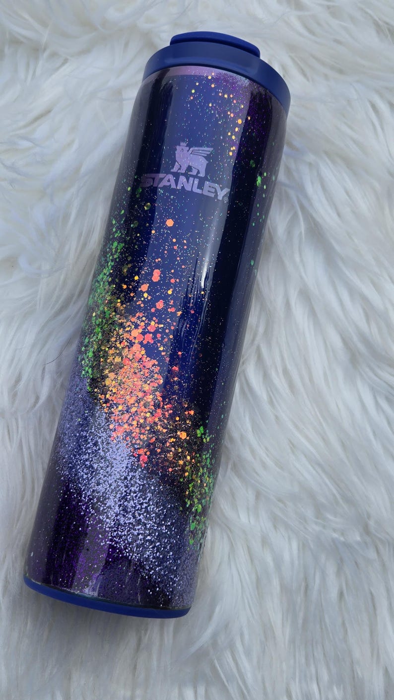 May include: A stainless steel Stanley tumbler with a navy blue base and lid. The tumbler is decorated with a galaxy-themed design, featuring colorful speckles of orange, green, and purple glitter. The Stanley logo is visible on the side.