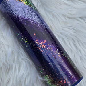May include: A cylindrical tumbler with a dark blue lid and base. The body of the tumbler is a gradient of dark blue to purple, speckled with iridescent glitter in shades of orange, green, and yellow. The tumbler sits on a white, fluffy surface.