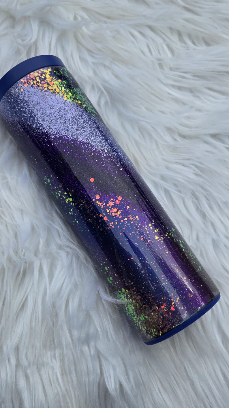 May include: A cylindrical tumbler with a dark blue lid and base. The body of the tumbler is a gradient of dark blue to purple, speckled with iridescent glitter in shades of orange, green, and yellow. The tumbler sits on a white, fluffy surface.