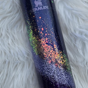 May include: A dark blue Stanley tumbler with a galaxy-inspired design. The tumbler features a speckled pattern of orange, green, and white glitter. The Stanley logo is visible on the side. The lid and base are dark blue.