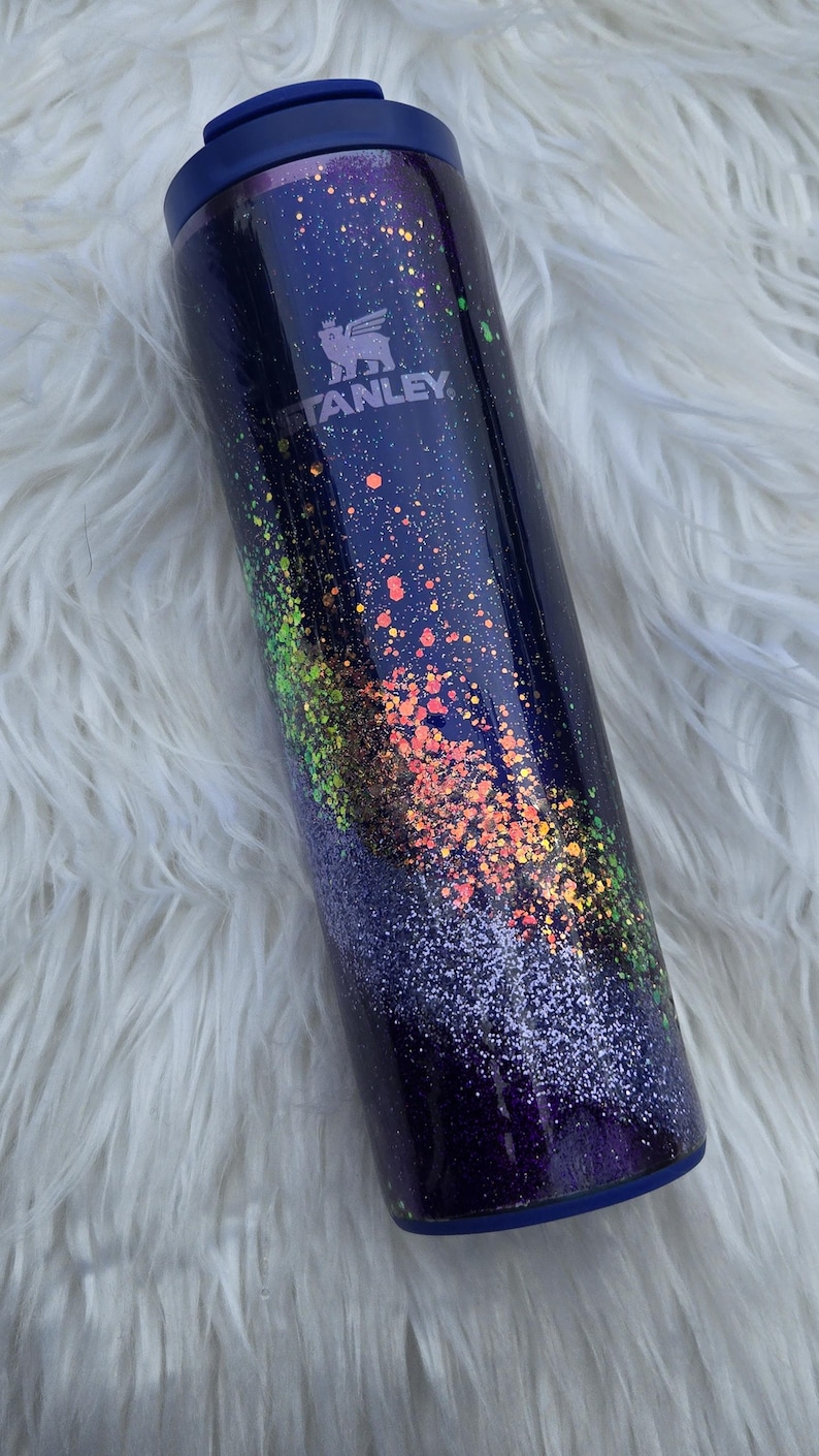 May include: A dark blue Stanley tumbler with a galaxy-inspired design. The tumbler features a speckled pattern of orange, green, and white glitter. The Stanley logo is visible on the side. The lid and base are dark blue.