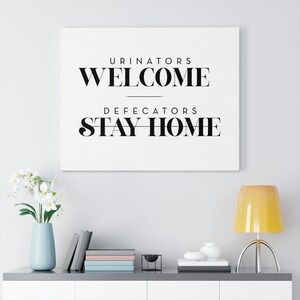 Puede incluir: Un cartel blanco de lona con texto negro que dice "Urinators Welcome Defecators Stay Home". El cartel está colgado en una pared blanca sobre un estante blanco con un jarrón de flores blancas, libros y una lámpara.
