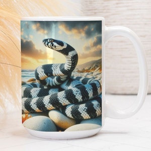 May include: White ceramic mug featuring a detailed illustration of a black and white banded snake coiled on a rocky beach. The background shows a sunset over the ocean. The mug has a large, curved handle.