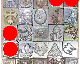 May include: A collection of square metal stamps featuring various embossed designs including a tent with the word CAMP, a cross, a lion, a giraffe, a dog, a goat, a ring, a Viking ship, a winged heart, a Celtic knot, and animal motifs.