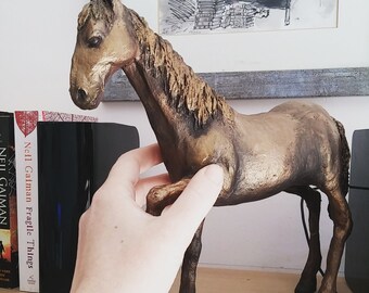 May include: A bronze-colored horse figurine with a flowing mane and tail. The horse is standing on a wooden surface.