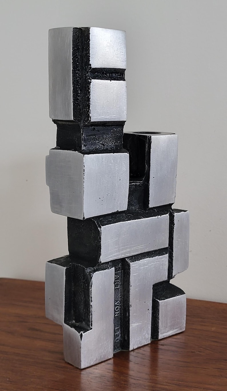 May include: A silver and black abstract sculpture made of stacked geometric shapes. The sculpture is signed 'ART VON LEO'.