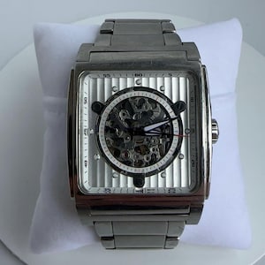 May include: A silver-toned, square-faced wristwatch with a skeleton dial, showcasing the inner workings. The watch has a silver band and a white face with black markings. The watch is displayed on a white pillow.