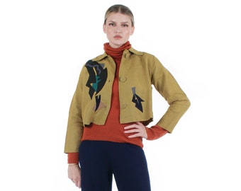 Puede incluir: Una mujer lleva una chaqueta color mostaza con diseños abstractos en negro y verde azulado, un jersey de cuello alto color óxido y pantalones de pierna ancha azul marino. El conjunto se completa con tacones beige. El fondo es blanco.