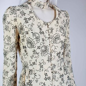 May include: A cream-colored dress with a black floral pattern and a high, open collar. The dress has long sleeves and a button-up front.