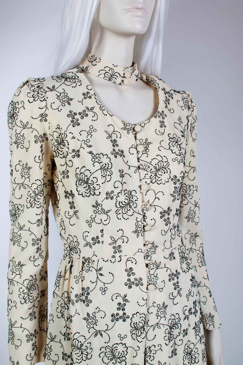May include: A cream-colored dress with a black floral pattern and a high, open collar. The dress has long sleeves and a button-up front.