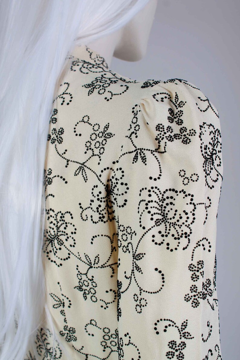 May include: A cream-colored blouse with a black floral pattern. The pattern is made up of small black dots that form flowers and vines.