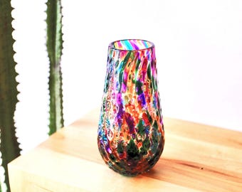 Könnte beinhalten: Eine farbenfrohe Glasvase mit einem gesprenkelten Muster in Rot-, Grün-, Lila- und Blautönen. Die Vase hat einen abgerundeten Boden und eine leicht ausgestellte Oberseite mit Regenbogenstreifen. Sie steht auf einer hellen Holzoberfläche.