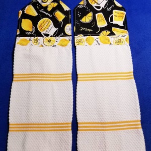 May include: Two white kitchen towels with yellow stripes. Each towel has a black and yellow lemon-themed fabric hanging loop attached. The loop on the left towel has the text "lemons" and the text "zesty" on the right loop.