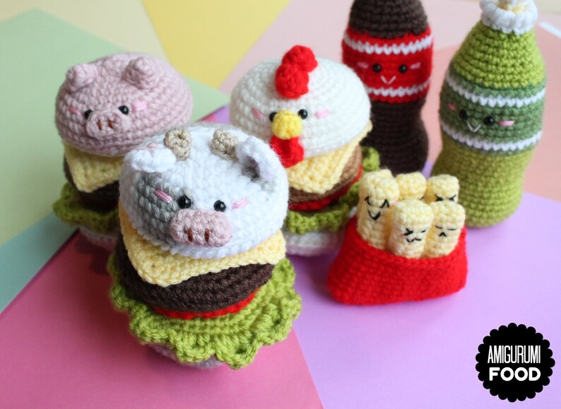 May include: A collection of crocheted amigurumi food items including two animal-themed burgers, a red container of french fries, and two beverage bottles. Each item features embroidered facial expressions. A black circular logo reads AMIGURUMI FOOD.