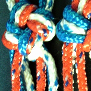 May include: Close-up of three strands of red, white, and blue braided rope. The rope is tied in a knot at the top and the strands are braided together in a complex pattern.