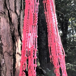 May include: A vibrant pink macrame plant hanger suspended from a tree branch. The handmade hanger holds a dark green pot with trailing plants. The macrame design features intricate knots and a tassel detail, adding a touch of bohemian style.