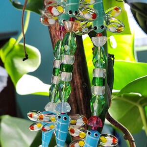 Può includere: Primo piano di ornamenti a forma di libellula in vetro colorato. Le libellule sono in tonalità blu e verde, con ali con accenti rossi, gialli e bianchi. Gli ornamenti sono infilati con perline trasparenti e verdi, creando un oggetto decorativo stravagante.