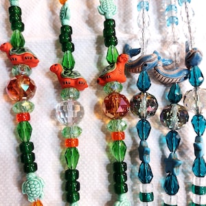 Puede incluir: Tres collares de cuentas con cuentas y dijes de diferentes colores. El primer collar tiene cuentas naranjas, verdes y turquesas con dijes de tortuga. El segundo collar tiene cuentas naranjas, verdes y turquesas con dijes de tortuga. El tercer collar tiene cuentas azules, blancas y verdes con dijes de ballena.