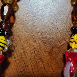 May include: Close-up of a beaded necklace featuring yellow and black bee charms, red flower charms, and various amber and clear beads. The necklace is set against a wooden background.
