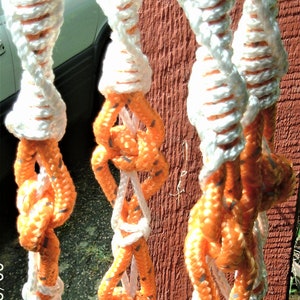 Puede incluir: Primer plano de una cuerda blanca y naranja con un diseño trenzado. La cuerda está anudada y cuelga de una superficie de madera.