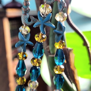 Peut inclure: Gros plan sur des bijoux de perles sur le thème des sirènes et des étoiles de mer. Les brins présentent des étoiles de mer bleues, des breloques de sirène turquoise et des perles à facettes dans des tons de jaune, transparent et turquoise. Les bijoux ont une esthétique côtière ou nautique.