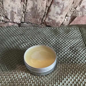 May include: A small, round, silver tin container filled with a light yellow balm or cream. The container is sitting on a green and white patterned surface.
