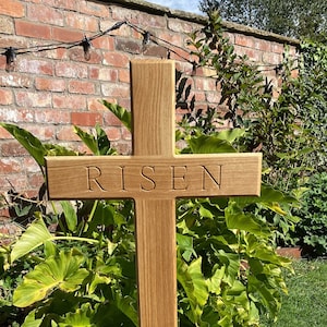 May include: A wooden cross with the word "RISEN" carved into it. The cross is standing on a wooden base and is in a garden setting.