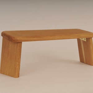 Puede incluir: Un banco de meditación de madera con un acabado natural. El banco tiene un diseño simple y rectangular con bordes redondeados y dos patas a cada lado. Las patas están ligeramente inclinadas hacia adentro.