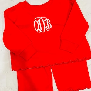 Puede incluir: Un conjunto de pijama rojo brillante con una camiseta de manga larga con cuello redondo y pantalones a juego. La parte superior tiene un diseño de monograma blanco. Los pantalones y la parte superior tienen un borde festoneado.