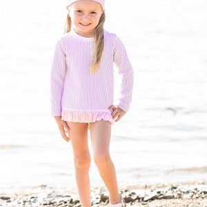 May include: A young girl wearing a pink and white striped long-sleeved rash guard with a pink polka dot headband. The rash guard has a ruffle trim at the bottom.