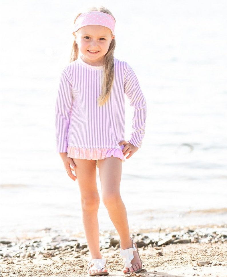 May include: A young girl wearing a pink and white striped long-sleeved rash guard with a pink polka dot headband. The rash guard has a ruffle trim at the bottom.