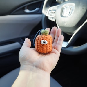 Op de afbeelding: Een kleine gehaakte pompoen met een groene steel en witte ogen. De pompoen is oranje en heeft een vriendelijke uitdrukking.
