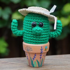 May include: A green crocheted cactus wearing a white straw hat with a white ribbon. The cactus is in a terracotta pot with a painted design of lavender flowers and bees.