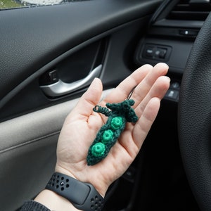 May include: A crocheted green pea pod keychain with three green peas inside. The keychain is held in a person's hand inside a car.