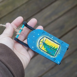 May include: A blue wooden sled ornament with a painted window scene of a green Christmas tree with red ornaments. The sled has a blue rope attached to it.